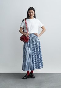 Femeie în picioare purtând un tricou alb, o fustă lungă albastru în carouri, șosete roșii, mocasini negri și având o geantă roșie pe umăr.