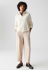 Pullover a costine crema con colletto a zip, abbinato a pantaloni beige a gamba larga e mocassini scuri. L'outfit presenta colori neutri e tessuti strutturati.