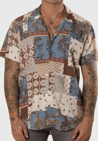 Camisa de manga corta con un diseño de patchwork que presenta varios patrones en azul, marrón, crema y rojo. El material parece ligero y tiene un ajuste relajado.
