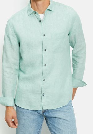 Mand iført en lysegrøn langærmet skjorte med knapper over en hvid undertrøje og lyseblå jeans, med ærmerne rullet op.