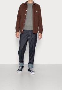 Brun skjorta med knappar, grå stickad tröja, mörk denimjeans med uppvikta manschetter och svarta högskaftade sneakers. Enkelt, avslappnat outfit.