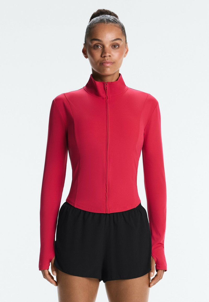 Femme aux cheveux attachés, portant une veste rouge zippée à manches longues et un short de sport noir, face à un arrière-plan uni.