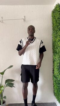 Witte kabelgebreide trui met zwarte strepen, gedragen over een zwart T-shirt. Gecombineerd met zwarte cargoshorts en zwarte sokken, tegen een groene achtergrond.