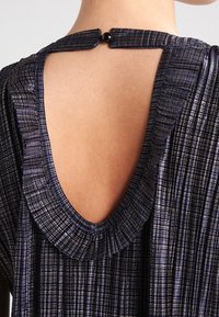 Black pleated dress features a round neckline, open back, and textured fabric with subtle metallic threads. Button closure at the neck.