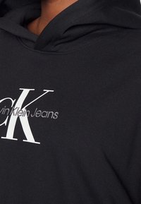Felpa nera con cappuccio realizzata in tessuto morbido, caratterizzata da un grande logo bianco con la scritta "Calvin Klein Jeans" che presenta una texture sottile e un design moderno.