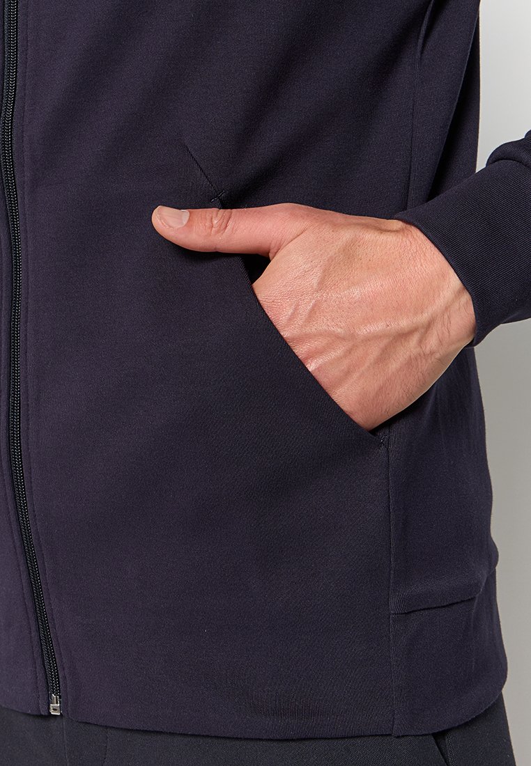 Dettaglio di una giacca blu navy con zip, caratterizzata da una tasca con una mano all'interno. Tessuto morbido e liscio con cuciture minime visibili.