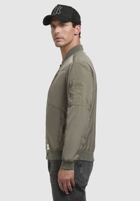 Licht olijfgroene bomberjack van nylon met ribgebreide boorden, een rits aan de voorkant en een utility pocket op de linker mouw. Paar donkere jeans.