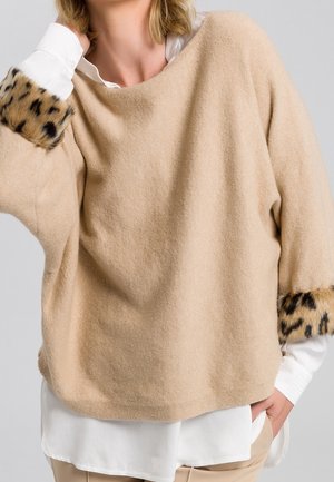 Donna che indossa un maglione beige con polsini in pelliccia leopardata sopra una camicia bianca e pantaloni beige, con la mano in tasca, su sfondo neutro.