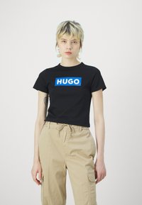 Juodas medvilnės marškinėliai su dideliu mėlynu stačiakampiu grafiniu elementu, ant kurio balta užrašas "HUGO". Derinama su bežinėmis kargo kelnių, kurios suteikia laisvą stilistiką.