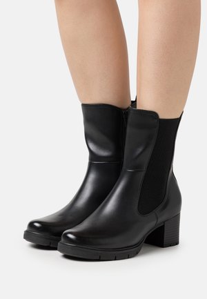 Bottes en cuir noir mi-mollet avec des talons bloc et des panneaux élastiques sur les côtés, portées sur des jambes nues, présentées sur un fond blanc.