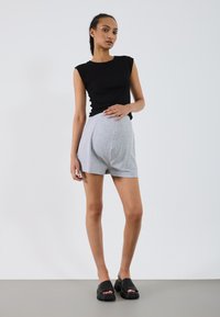 Top aderente senza maniche nero, pantaloni corti grigi con design plissettato e scarpe slip-on con piattaforma nere, presentate su uno sfondo semplice.