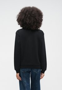 Άτομο με σγουρά μαλλιά, φορώντας απλό μαύρο φούτερ και μπλε τζιν, στέκεται με την πλάτη προς τα εμπρός σε ένα απλό λευκό φόντο.