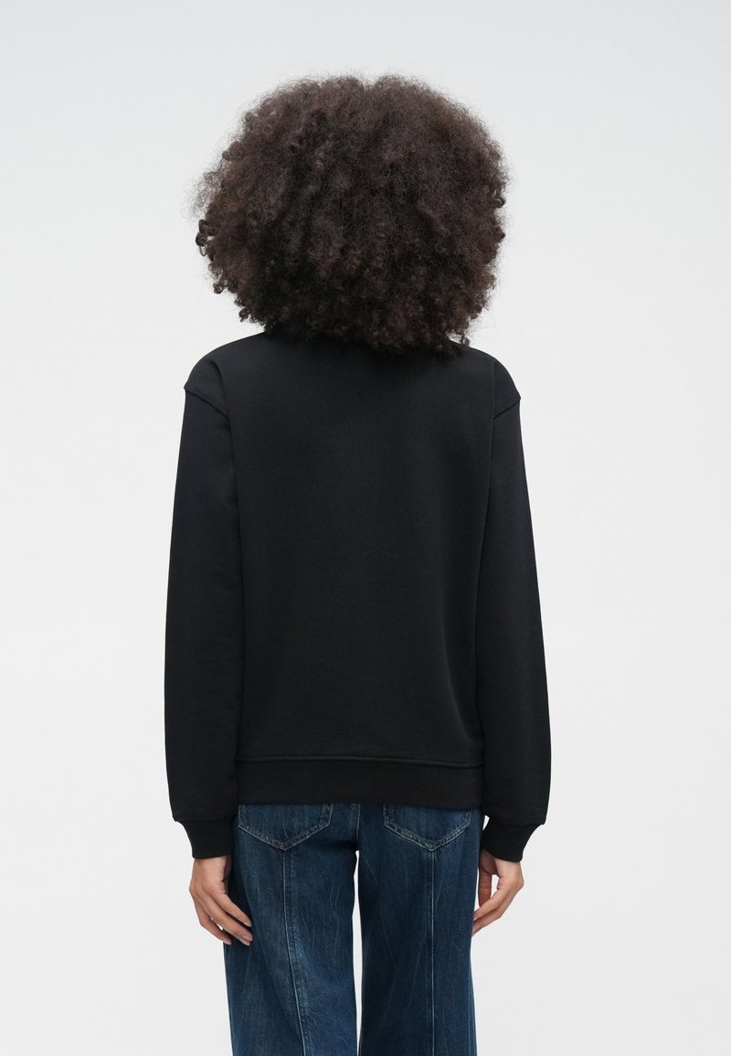 Persona con capelli ricci che indossa una felpa nera semplice e jeans blu, in piedi di schiena su uno sfondo bianco uniforme.