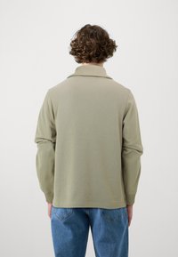 Grønn strikket genser med ribbestrikket krage, lange ermer og sidesplitter. Har en avslappet passform og glatt tekstur, stylet med blå jeans.