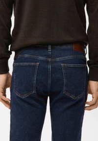 Mann trägt dunkelblaue Jeans und ein schwarzes Langarmshirt, von hinten gezeigt, Hände entspannt an den Seiten.