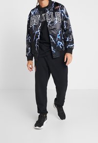 Mustaa satiinia oleva bomber-takki, jossa on sinisiä salaman kuva-aiheita ja "BULLS"-tekstiä, yhdistetty mustiin verkkareihin ja mustiin lenkkareihin.