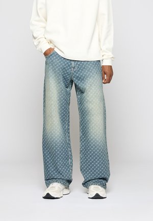 Homme portant un jean large bleu clair avec des imprimés blancs, des baskets blanches et une chemise blanche à manches longues, debout devant un fond uni.