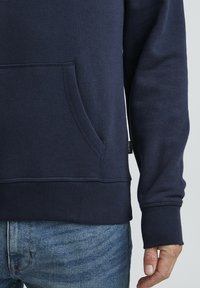 Felpa blu navy con tasca anteriore, polsini e fondo a costine, realizzata in tessuto morbido con una texture liscia.