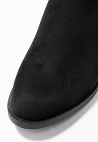 Svart mocka ankelsko med rund tå, jämn yta och en gummisula med räfflad kant. Närbild visar materialets detalj.