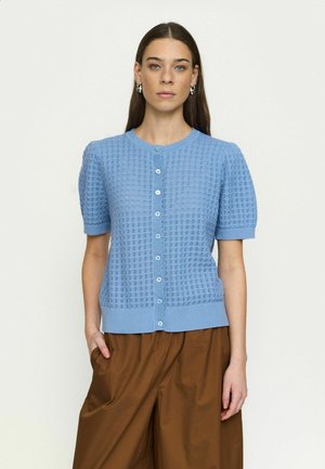 Hosszú barna hajú nő világoskék, gombos, rövid ujjú kötött felsőt és barna, bő szárú nadrágot visel, kezét az egyik zsebében tartva áll.