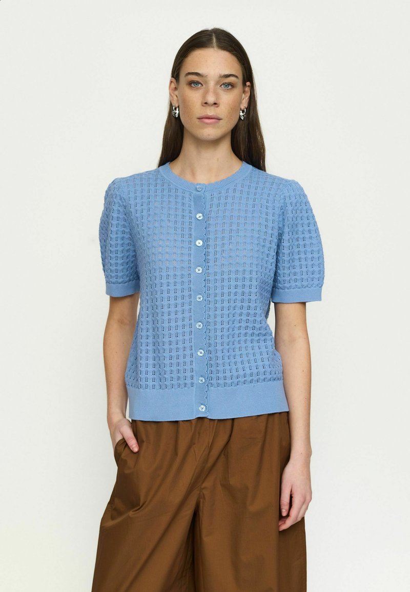 Moteris su ilgais rudais plaukais vilki šviesiai mėlyną trumpomis rankovėmis megztą palaidinę su sagomis ir rudus, plačias kelnes, stovinti viena ranka kišenėje.