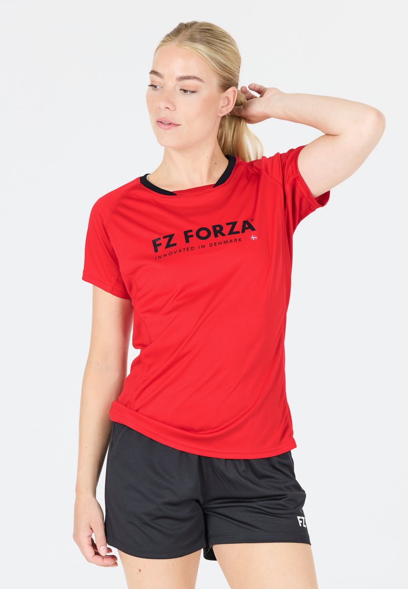 Kvinna klädd i röd FZ Forza sporttröja och svarta shorts, som justerar sitt hår med ena handen mot en enkel vit bakgrund.