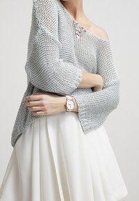 Donna che indossa un maglione grigio a spalle scoperte e una gonna bianca, con un orologio al polso sinistro e una collana visibile vicino alla clavicola.