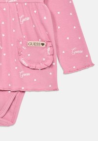 Rosa ribbet heldrakt med hvite hjertepatter, lange ermer og volanglomme. "Guess"-logoen vises i skrifttype på stoffet og lommen.
