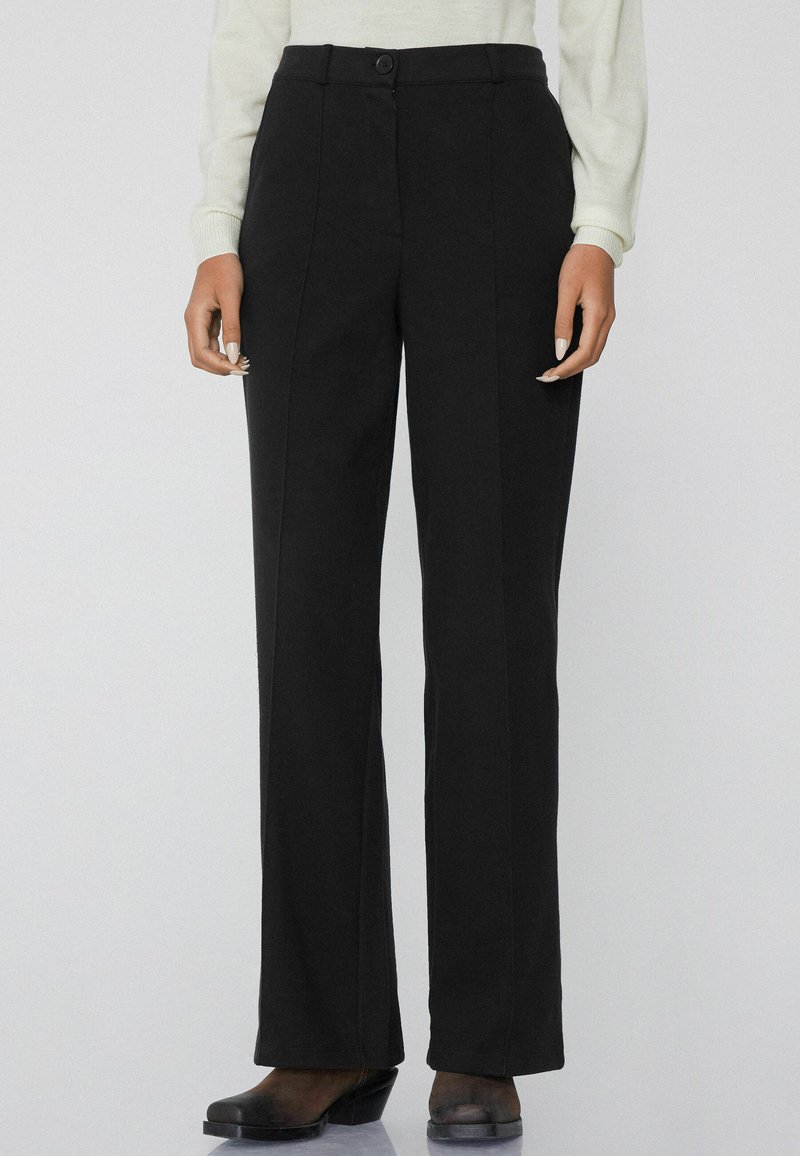 Zwarte wijde broek van gladde stof met een hoge taille, knoopsluiting en voornaaddetails. Gecombineerd met zwarte hakken.