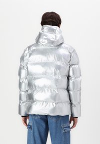 Giacca imbottita argento con finitura lucida, dotata di baffles, cappuccio e polsini elasticizzati. Indossata sopra pantaloni cargo blu.