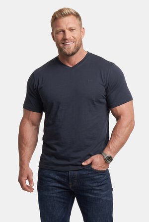  muskularny mężczyzna o blond włosach i brodzie uśmiecha się, nosząc ciemnogranatowy t-shirt z dekoltem w serek i niebieskie dżinsy na jednolitym tle.