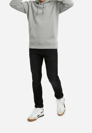 Persona vistiendo una sudadera con capucha gris claro, jeans ajustados negros y zapatillas blancas con suelas marrones, de pie con los brazos levantados.