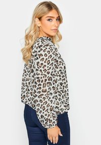 Blusa traslucida con maniche lunghe, caratterizzata da uno sfondo beige e fantasie a stampa leopardo nero-marrone. Polsini elasticizzati e scollatura arricciata.