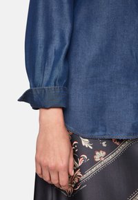 Denim blouse with long, gathered sleeves and a buttoned cuff; features blue color, stitched seams, and a smooth texture.