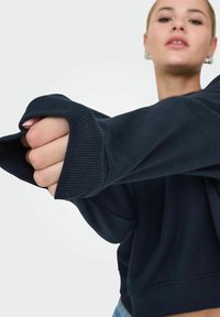 Sweatshirt noir surdimensionné avec des poignets côtelés. Le matériau semble doux et texturé. Détail d'une main tenant le tissu est visible.