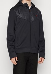 Black hoodie with a glossy, patterned upper chest section, zip closure, and side pockets, made from soft, textured fabric with ribbed cuffs.