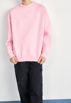 Personne portant un sweat-shirt New Balance rose clair et un pantalon cargo noir ample, debout contre un mur clair uni.