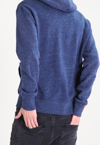 Sudadera azul marino con una tela texturizada y moteada, que cuenta con una capucha ajustable, puños y dobladillo de canalé. Lleva puesta sobre pantalones negros con bolsillos traseros.