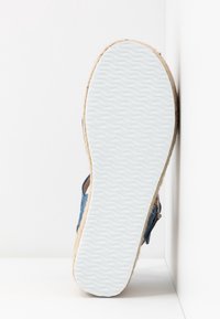Semelle en caoutchouc blanche avec motif ondulé sur une sandale espadrille bleue reposant verticalement contre une surface blanche.