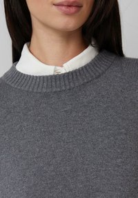Vrouw in een wit gestructureerd coltrui met daaronder een grijze gebreide trui met ronde hals, waarbij haar lippen, nek en bovenborst zichtbaar zijn.