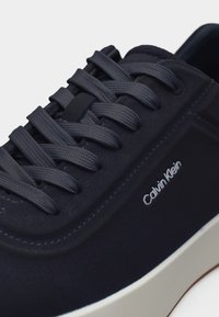 Αθλητικό παπούτσι Calvin Klein σε ναυτικό μπλε, με υφασμένα κορδόνια, ραμμένες λεπτομέρειες και λευκή σόλα, σε κοντινή όψη από το πλάι και πάνω.