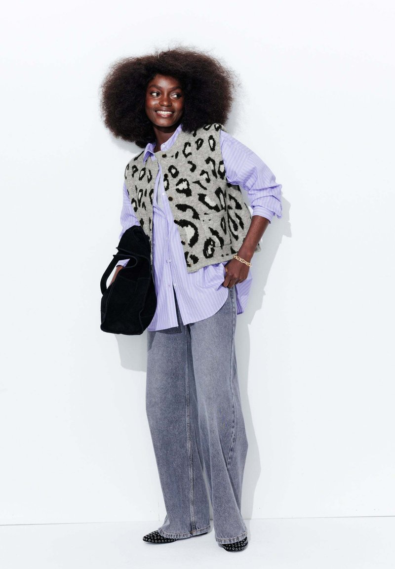 Un gilet en fausse fourrure imprimé léopard porté sur une chemise rayée lilas, associé à un jean gris large et des ballerines noires pointues, tenant un sac fourre-tout noir.