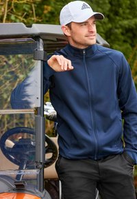 Chaqueta navy con cremallera y lados texturizados, pantalones negros, llevada con una gorra blanca con un logo negro, de pie junto a un carrito de golf.