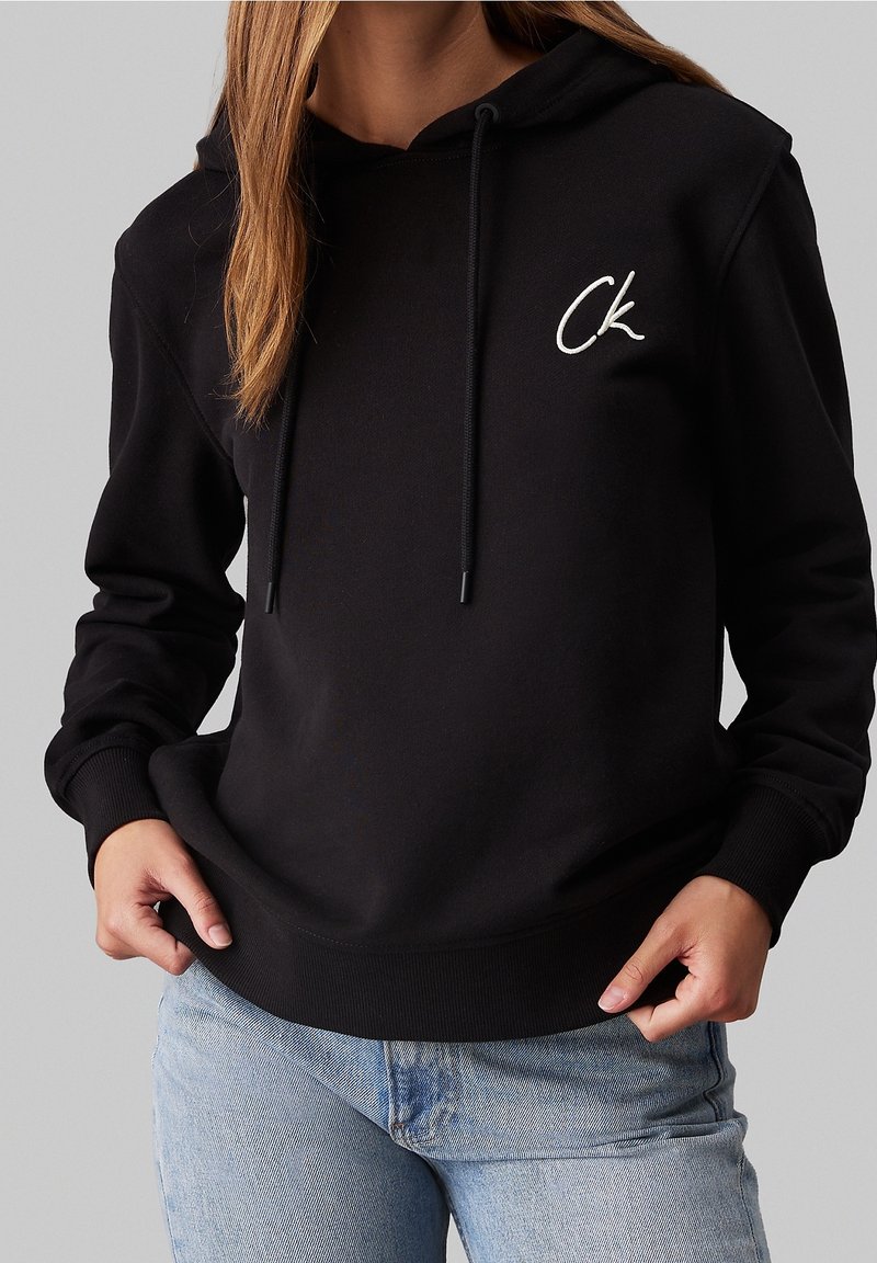Sweat à capuche noir avec logo blanc "Ck" sur la poitrine, porté par une personne aux cheveux longs, associé à un jean bleu clair sur fond gris uni.