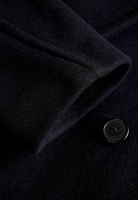 Jūras zila vilnas pieta ar maigām tekstūrām, ar melna pogas detaļu un salocītu apkakli. Tuvplāns pie atloku zonas.