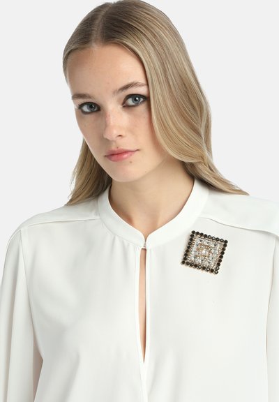Bluză albă cu decolteu tip cheie, decorată cu un broșă pătrată cu bijuterii, având cristale negre și clare care accentuează zona pieptului.