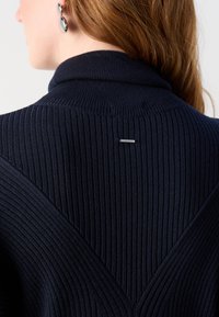 Suéter de punto de color marino con cuello alto, que presenta un patrón texturizado y un pequeño detalle de hardware plateado en la parte posterior.