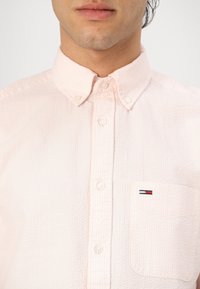 Világos rózsaszín gombos ing, texturált anyagból, mellzsebbel és egy kis logóval, valamint klasszikus gallérral.