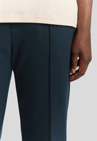 Marineblaue Sporthose aus weichem Stoff, mit schlanker Passform und vertikalen Nahtdetails, kombiniert mit einem hellbeigen T-Shirt.