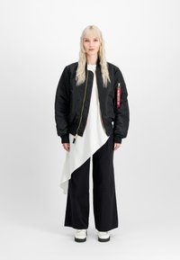 Jachetă bomber neagră cu un fermoar aurie, asociată cu un top alb asimetric și pantaloni negri largi, purtată peste încălțări cu platformă albe.